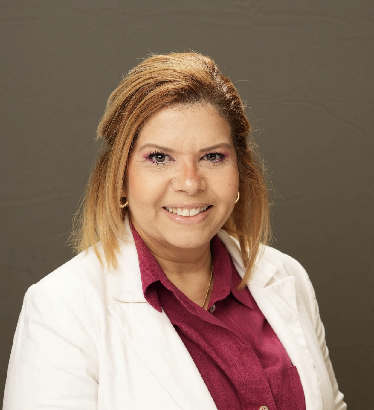 Ninoska Eurea, a woman with shoulder-length light brown hair, wears a white blazer over a magenta shirt and smiles at the camera against a plain gray background.